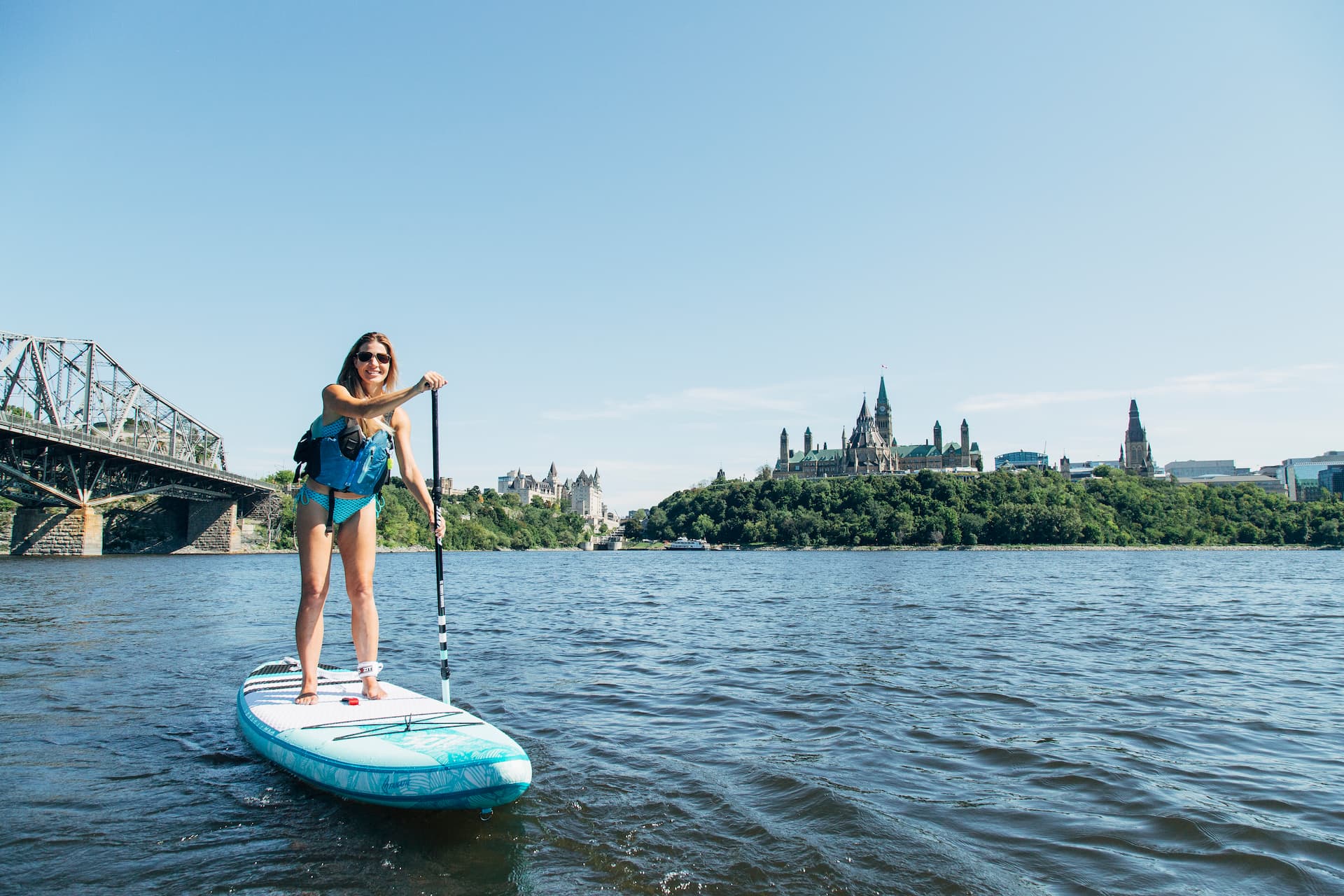 A person paddleboarding on a river with a cityscape and bridge visible in the background under a clear sky.