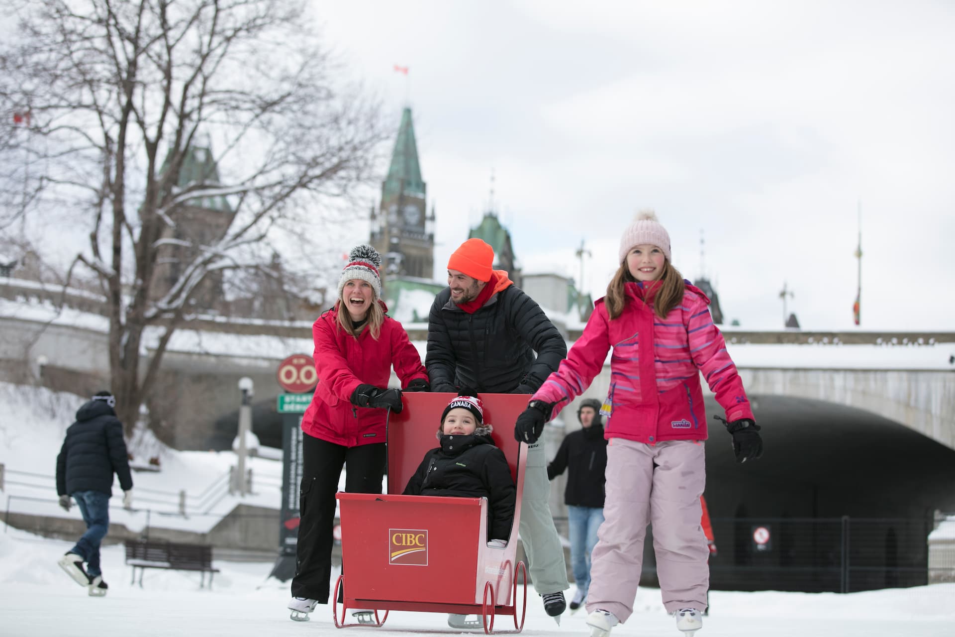 A family pushes a child in a red sled on an outdoor ice rink in winter, with historic buildings in the background.