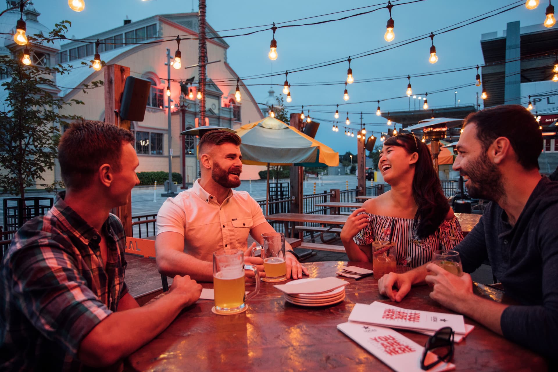 Four people sit at an outdoor table with drinks, laughing and talking under string lights in the evening.