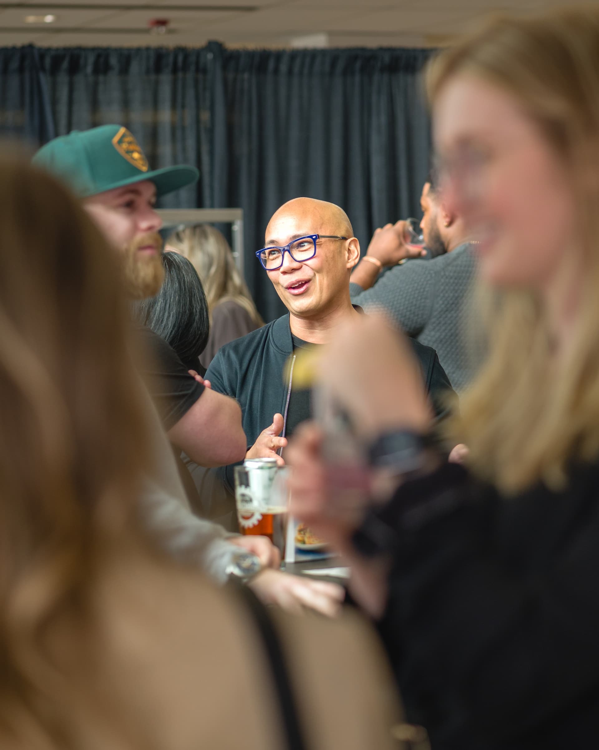 A man with glasses smiles and talks to others at a social event with drinks in hand.