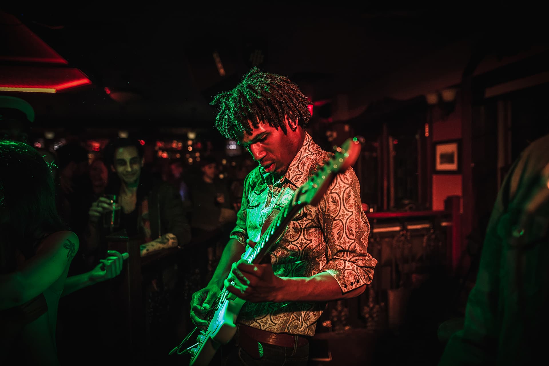 A musician plays an electric guitar on stage under red and green lighting with a crowd watching in the background.