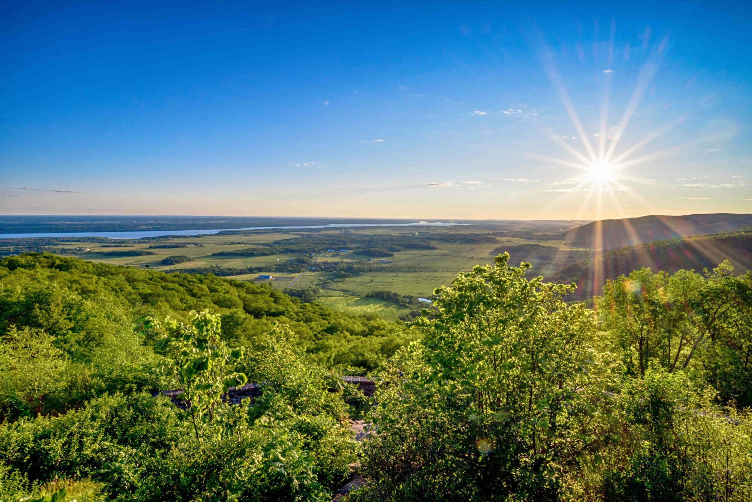 The sun shines brightly over a green, rolling landscape with distant water under a clear blue sky.