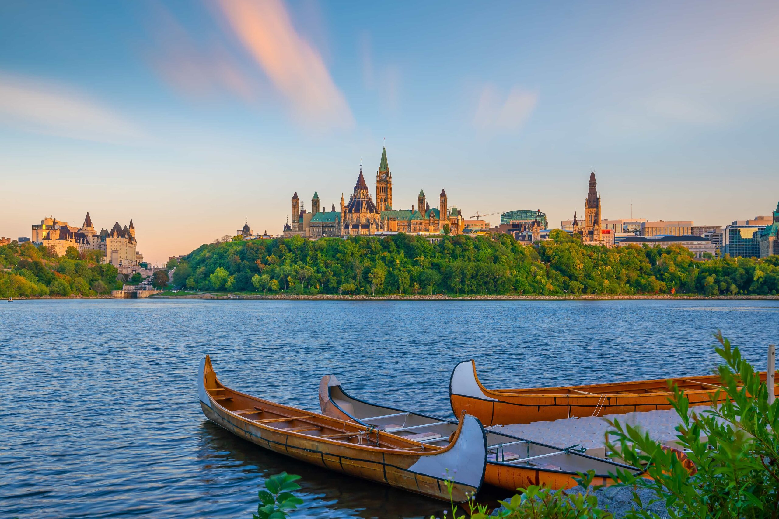 Three canoes on a river with Ottawa’s Parliament buildings atop a green hill in the background at sunset.
